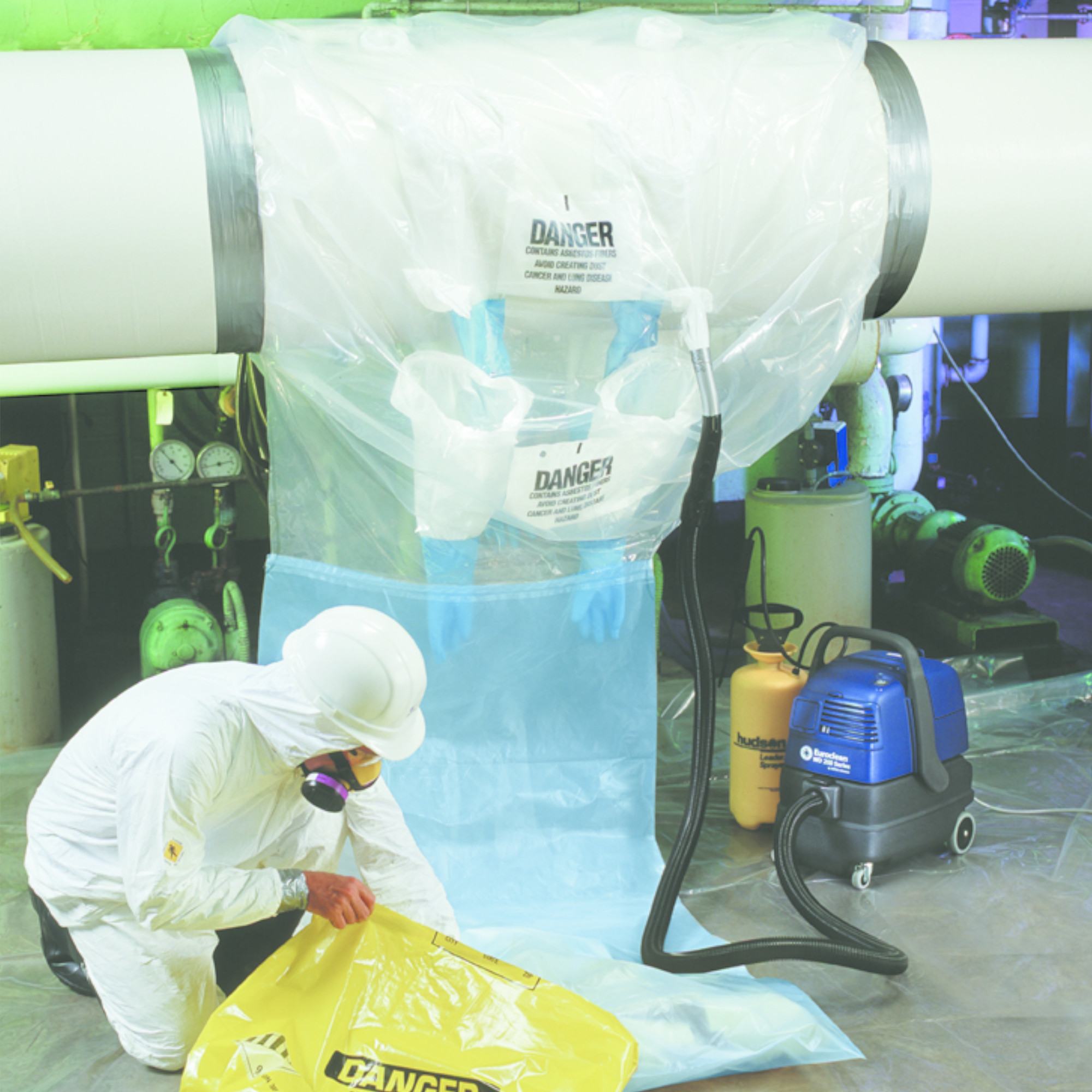 Eine Person in Schutzkleidung und Helm beugt sich über einen gelben Gefahrensack in einem industriellen Raum. Im Hintergrund befindet sich eine transparente Abdeckung an einem Rohr, auf der Warnhinweise sowie ein Absauggerät zu sehen sind. Die Umgebung deutet auf Arbeiten mit gefährlichen Stoffen hin.